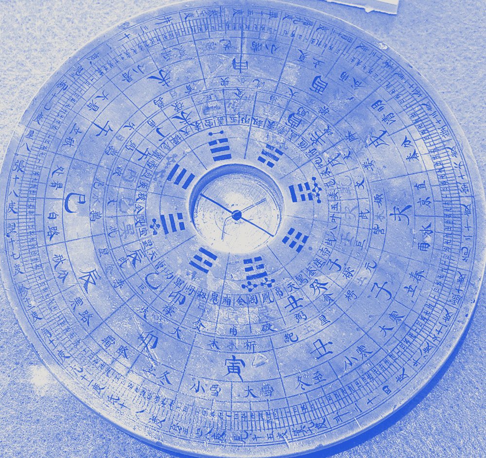 A wooden geomantic compass, its rings of characters and trigrams around a magnetic needle, printed in blue